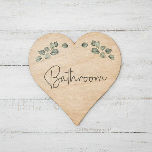 A heart-shaped wooden bathroom sign with a natural light wood grain finish, featuring green eucalyptus leaf illustrations and the word 'Bathroom' written in black cursive script, shown on a plain white background.