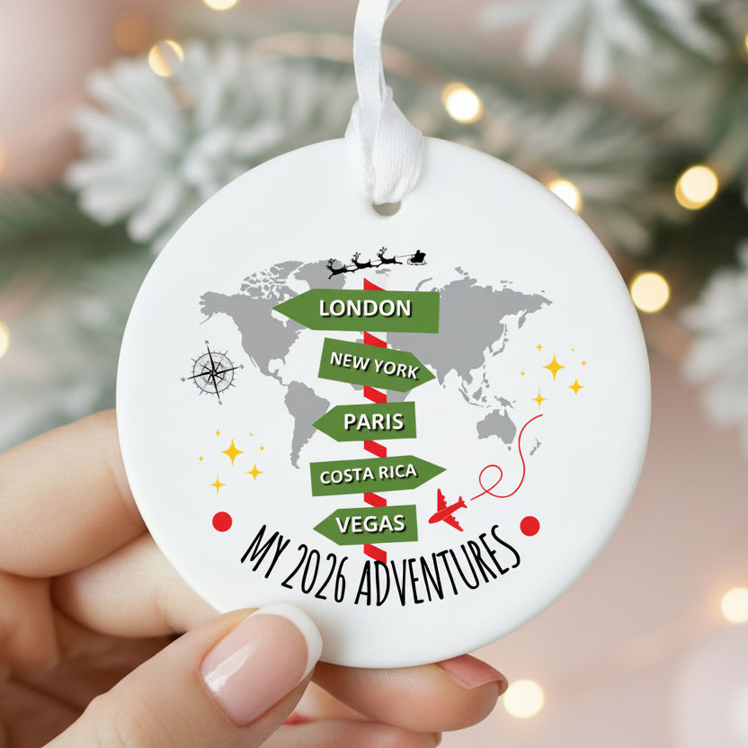 A round white Christmas ornament held by hands, featuring a gray world map with green directional flight-path style labels for London, New York, Paris, Costa Rica, and Vegas, plus the text 'MY 2026 ADVENTURES' and a white hanging loop.