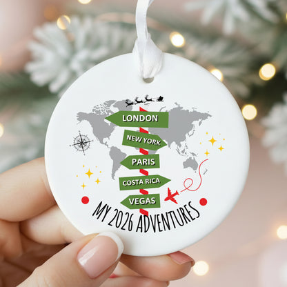A round white Christmas ornament held by hands, featuring a gray world map with green directional flight-path style labels for London, New York, Paris, Costa Rica, and Vegas, plus the text 'MY 2026 ADVENTURES' and a white hanging loop.
