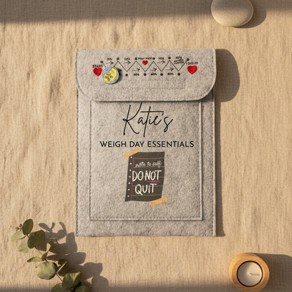 A gray felt pouch labeled 'Katie's WEIGH DAY ESSENTIALS' with a flap closure, printed weight-chart graphics, and a pocket reading 'write to self DO NOT QUIT', shown on a white desk with a keyboard corner and binder clip nearby.